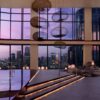 Luxurious indoor pool area with a city skyline sunset, illuminated steps, and lounge chairs along the right, featuring circular hanging lamps overhead and large windows framing the urban view.