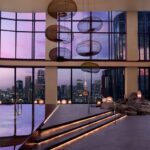 Luxurious indoor pool area with a city skyline sunset, illuminated steps, and lounge chairs along the right, featuring circular hanging lamps overhead and large windows framing the urban view.