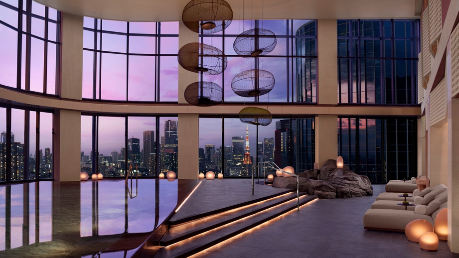 Luxurious indoor pool area with a city skyline sunset, illuminated steps, and lounge chairs along the right, featuring circular hanging lamps overhead and large windows framing the urban view.