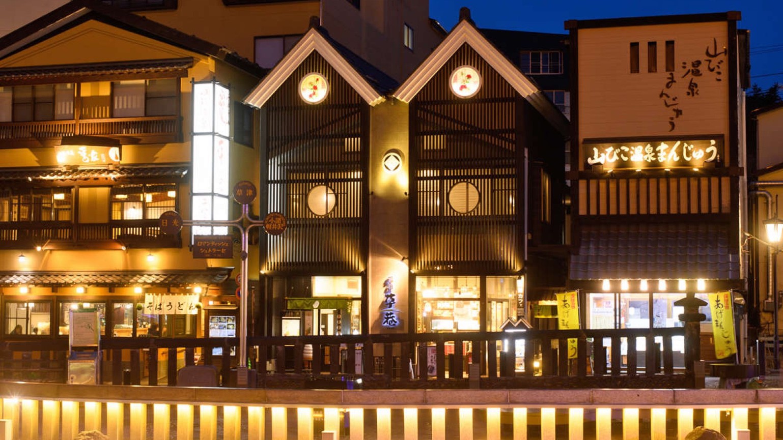 Nighttime street scene in Japan with warmly lit wooden storefronts and lanterns along a canal railing, signs in Japanese.