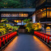 Red-painted pedestrian bridge at dusk leading to a traditional Japanese-style building with warm illuminated windows and lush greenery.