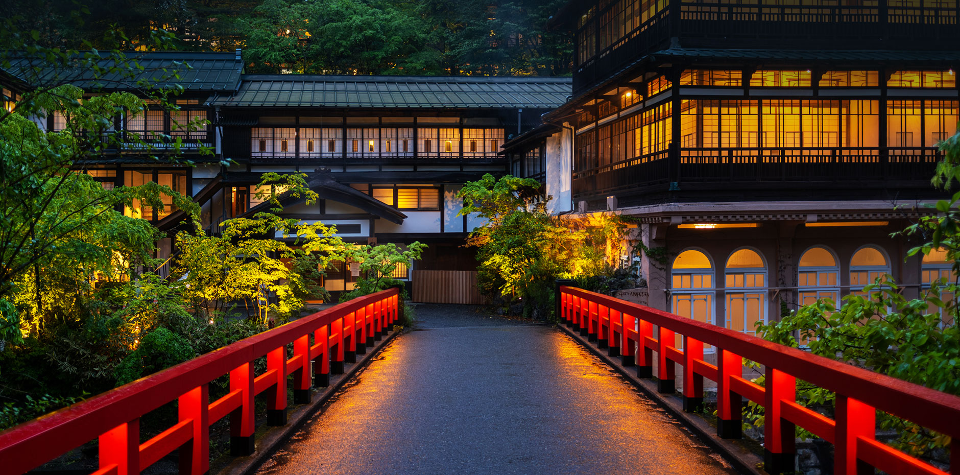 Red-painted pedestrian bridge at dusk leading to a traditional Japanese-style building with warm illuminated windows and lush greenery.