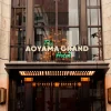 Hotel entrance with illuminated sign reading 'The AOYAMA GRAND Hotel' above glass doors, flanked by greenery on the building façade.