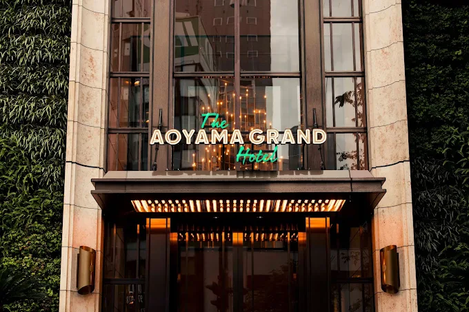 Hotel entrance with illuminated sign reading 'The AOYAMA GRAND Hotel' above glass doors, flanked by greenery on the building façade.