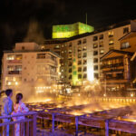 Couple in yukata overlook steaming onsen pools at night in a warmly lit town scene.