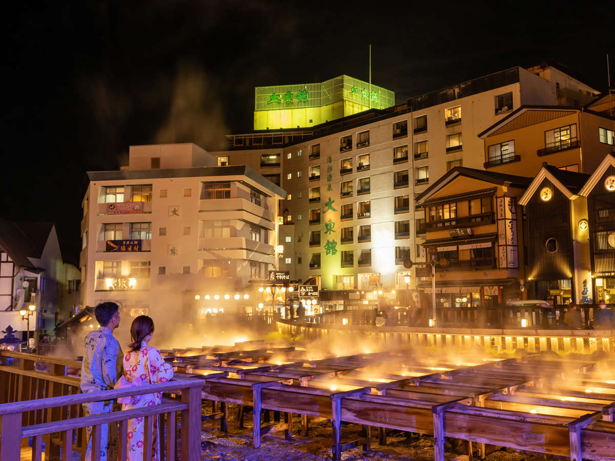 Couple in yukata overlook steaming onsen pools at night in a warmly lit town scene.
