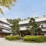 Traditional Japanese-style building with white walls and curved tiled roof, surrounded by trimmed shrubs and trees against a blue sky