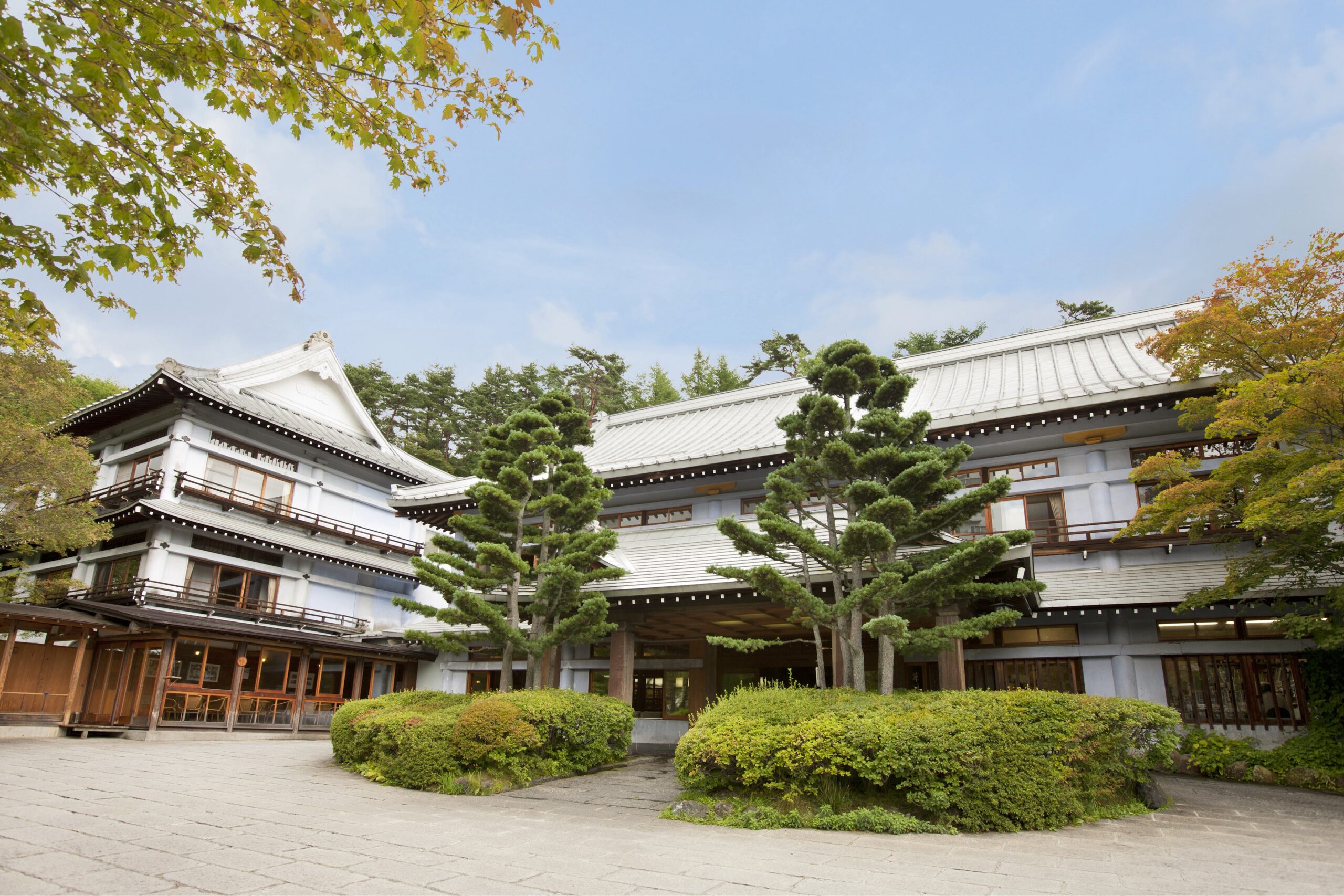 Traditional Japanese-style building with white walls and curved tiled roof, surrounded by trimmed shrubs and trees against a blue sky